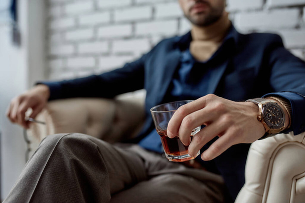 Selective focus of glass with cognac or whiskey in hands of partial blurred IT seller sit and rest in armchair in office. Modern successful male lifestyle. Man wearing formal wear and wristwatch - Valokuva, kuva