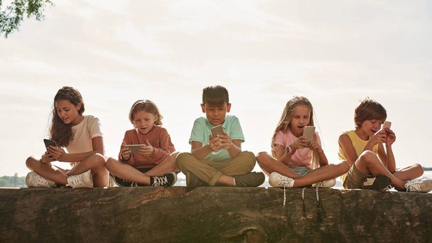感情的な多民族の子供たちのグループを使用して、屋外の川の海岸にログオンしてスマートフォンを見て。世代アルファの男の子と女の子。麻薬中毒だ。子供時代の生活。夏の晴れた日 - 写真・画像