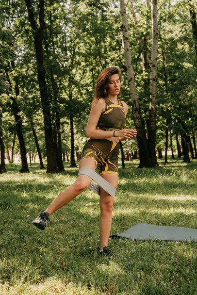公園や森の中でフィットネス用の伸縮性のあるスポーツをする少女です。ヨガマットに緑のスーツを着て - 写真・画像