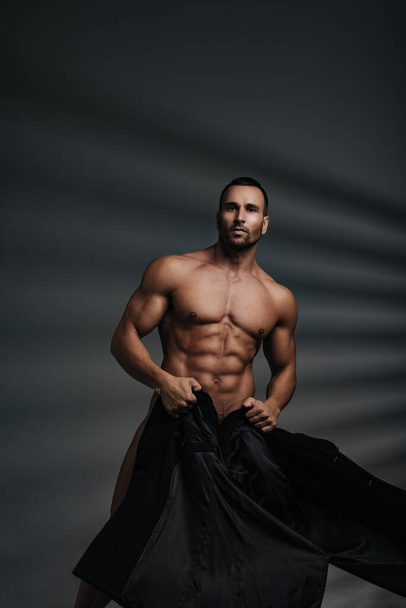 Gebräunter und muskulöser sexy nackter Mann mit schwarzem Mantel, der seinen Körper bedeckt. Kunst nackte Mode männliches Modell. - Foto, Bild
