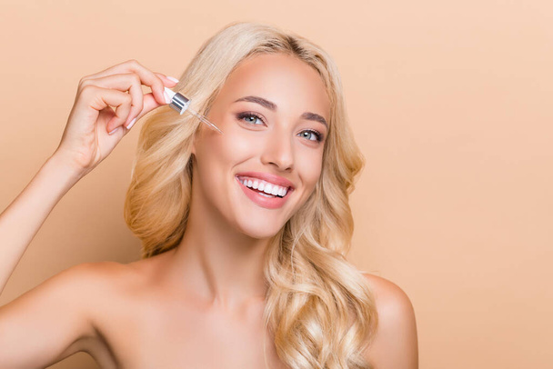 Photo of pretty positive girl with bare shoulders use essence on forehead with pipette isolated on beige color background. - Фото, изображение