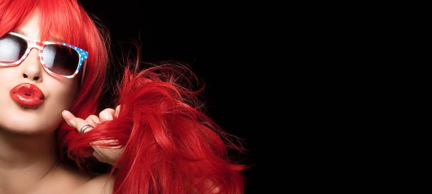 Beautiful sexy woman with long red hair wearing American flag sunglasses pouting her plump lips at the camera in a sensuous gesture. Beauty portrait isolated on black background - Foto, afbeelding