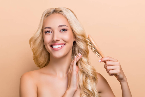 Photo of lovely positive female comb her amazing long curly silky hair with brush isolated on beige color background. - Zdjęcie, obraz