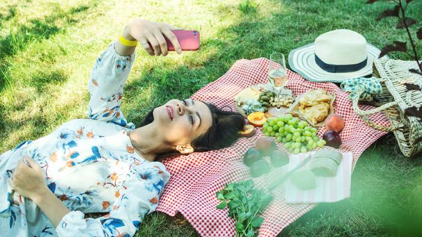 Kırsal kesimdeki bir çayırda piknik yaparken selfie çeken bir kadın. Güneşli bir günde battaniyeye uzanmak. - Fotoğraf, Görsel