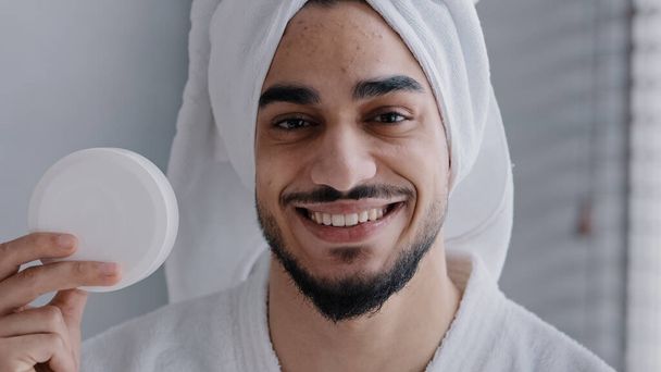 Arabian hispanic indian man with towel on head holding jar of cream for face laugh looking at camera recommend natural male cosmetic in bathroom advertise facial skin care moisture treatment hydration - Foto, imagen