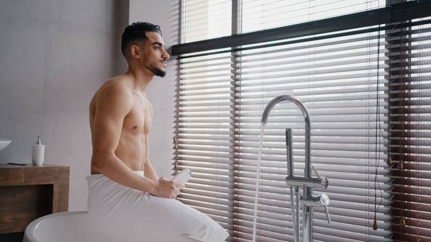 Morning bath, Arab Indian Spaniard naked sexy bearded man with bath towel on hips sits in bathroom looking at window holding shower gel natural male cosmetic morning refreshment hygiene procedure - 写真・画像