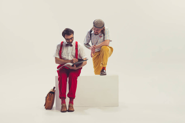 Fashion designer. Two cheerfull dudes, young men in old-school fashioned attire reading magazine isolated on white background. Artists in motion. Concept of culture, art, music, style, ad - 写真・画像