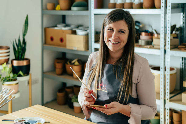 Retrato de feliz mujer de negocios en delantal pintando un corazón de arcilla en su escritorio de taller - Foto, Imagen