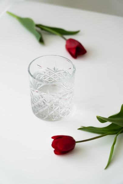 vidrio con agua pura cerca de tulipanes rojos frescos en la superficie blanca y fondo borroso - Foto, Imagen