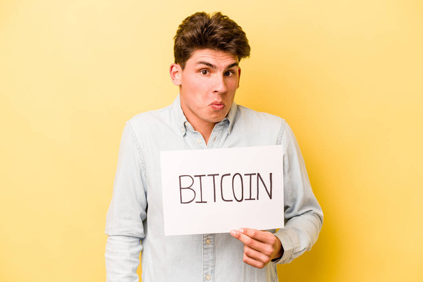 Giovane uomo caucasico in possesso di cartello bitcoin isolato su sfondo giallo spalle scrollate e occhi aperti confusi. - Foto, immagini