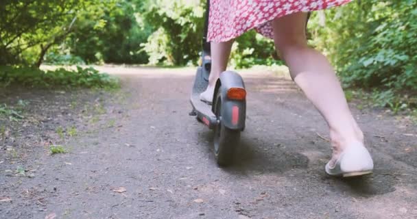 Mujer en vestido comienza a moverse en scooter eléctrico a lo largo del camino en el parque. Arranca el suelo con el pie. Vista trasera, primavera verano, día, árboles verdes a través del follaje el sol brilla. Alto. - Metraje, vídeo