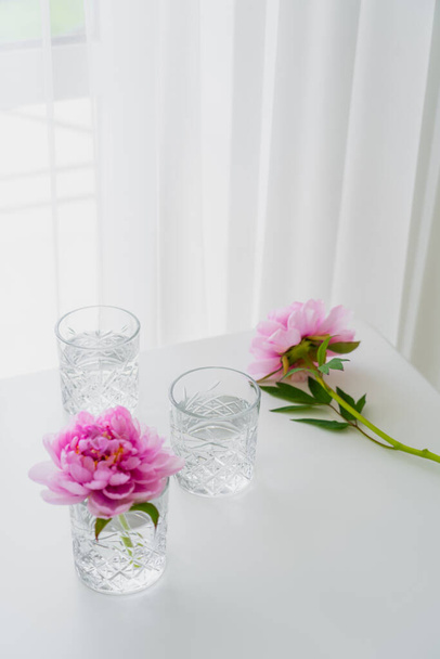 transparent faceted glasses and pink peonies on white tabletop near curtain - Fotó, kép