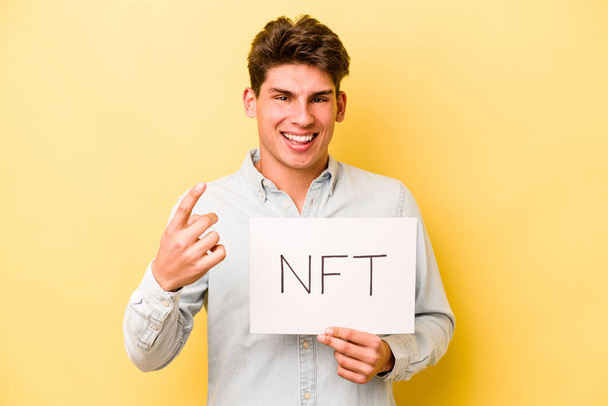 Νεαρός καυκάσιος που κρατάει την πινακίδα του NFT απομονωμένη σε κίτρινο φόντο δείχνοντάς σε με το δάχτυλο σαν να σε προσκαλεί να έρθεις πιο κοντά.. - Φωτογραφία, εικόνα
