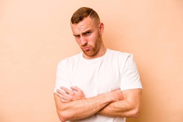 Young caucasian man isolated on beige background frowning face in displeasure, keeps arms folded. - Foto, imagen