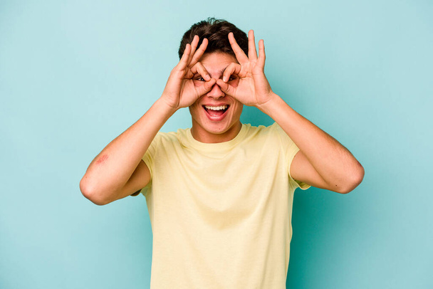 Giovane uomo caucasico isolato su sfondo blu mostrando segno ok sopra gli occhi - Foto, immagini