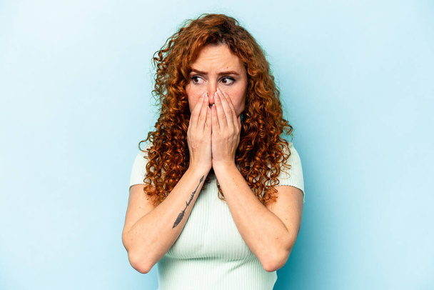 Young ginger caucasian woman isolated on blue background covering mouth with hands looking worried. - Foto, Imagen