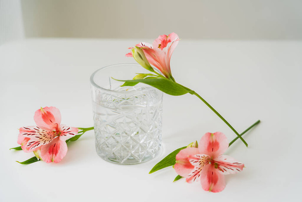 цветы альстромерии рядом с граненым стеклом с чистой водой на белой поверхности и сером фоне - Фото, изображение