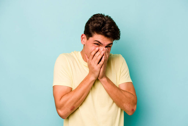 Jonge blanke man geïsoleerd op blauwe achtergrond lachen over iets, bedekken mond met handen. - Foto, afbeelding