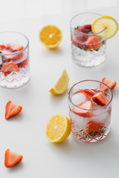 faceted glasses with fruit tonic drink near chopped strawberries and lemons on white tabletop - Foto, Bild