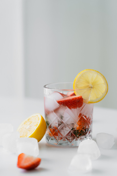 vidro de bebida tônica gelada com morangos picados perto de limão cortado na superfície branca e fundo cinza - Foto, Imagem