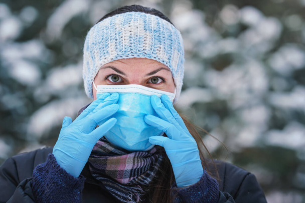 Young woman in winter jacket with blue disposable single use face virus mask and protective gloves, blurred snow covered trees background, closeup detail - Fotografie, Obrázek