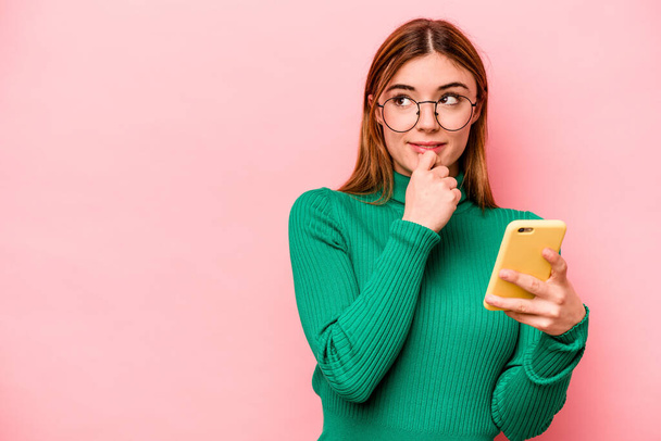 Young caucasian woman holding mobile phone isolated on pink background relaxed thinking about something looking at a copy space. - Foto, afbeelding