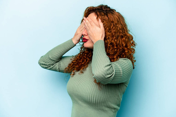 Νεαρή κοκκινομάλλα καυκάσια γυναίκα απομονωμένη σε μπλε φόντο φοβάται να καλύψει τα μάτια με τα χέρια. - Φωτογραφία, εικόνα