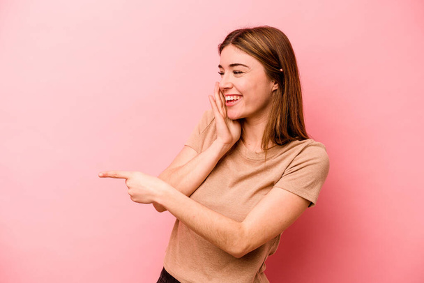 Young caucasian woman isolated on pink background saying a gossip, pointing to side reporting something. - Foto, afbeelding
