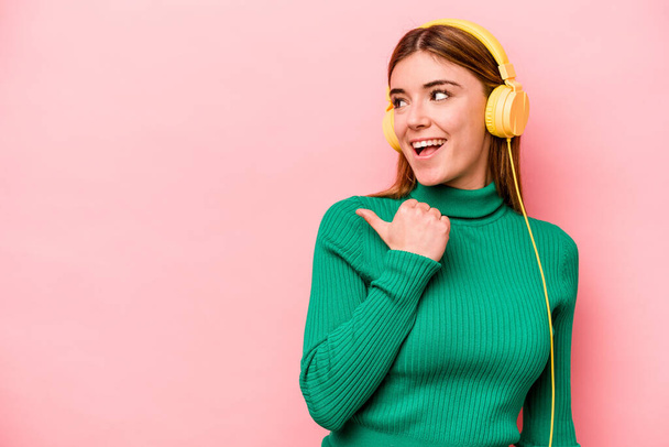 Young caucasian woman listening to music isolated on pink background points with thumb finger away, laughing and carefree. - Valokuva, kuva