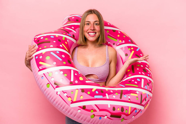 Mujer caucásica joven sosteniendo un colchón de aire aislado sobre fondo rosa mostrando un espacio de copia en una palma y sosteniendo otra mano en la cintura. - Foto, imagen