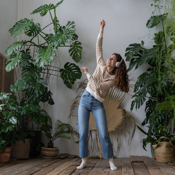 Carefree young woman with long hair in wireless headphones listening to music with pleasure, enjoying the moment, relaxing dancing on wooden floor in cozy home garden with monstera and tropical plants - Foto, Imagen