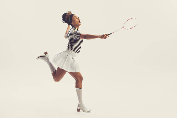 Leisure activities. Young happy beautiful girl in retro, 70s, 80s fashion style clothes, vintage outfit playing badminton isolated on white background. Concept of culture, art, music, fashion style. - Φωτογραφία, εικόνα