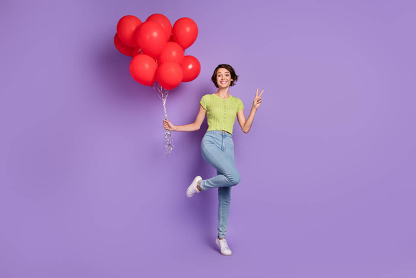 Photo of attractive beautiful young lady wear green t-shirt showing v-sign holding red balloons isolated violet color background. - 写真・画像