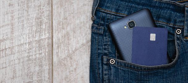 Bankpas en smartphone in spijkerbroekzak op lichte houten achtergrond, kopieerruimte links. - Foto, afbeelding