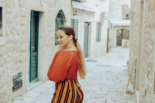 Beautiful stylish girl with long earrings, wearing an orange suit looks at the camera. Traveling around Europe Fashionable woman smiling in the old town and posing.  - Fotó, kép