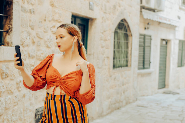 Woman in the old city and posing. Beautiful stylish girl with a smartphone in her hands, makes a photo or video, travel blogger. Traveling around Europe - Photo, Image
