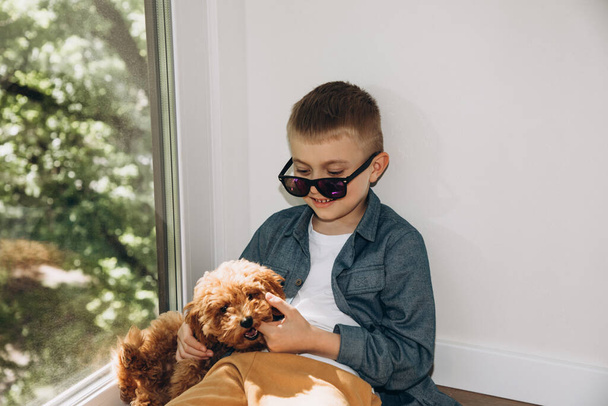 Χαρούμενο αγόρι 6-7 ετών με σκούρα γυαλιά ηλίου να κάθεται στο πάτωμα παίζοντας με ένα σκύλο - Φωτογραφία, εικόνα