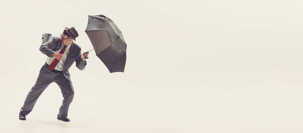 Bad weather. Portrait of young man dressed in 50s, 60s style fights the wind with umbrella isolated on white background. Autumn fashion collection. Concept of culture, art, wellbeing, beauty and ad. - Фото, зображення