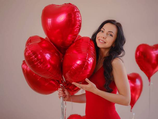 Krásná šťastná mladá žena v červených šatech. sváteční večírek. Radostný model pózující s červeným srdcem ve tvaru balónů, baví, slaví Valentýna. Symbol lásky - Fotografie, Obrázek