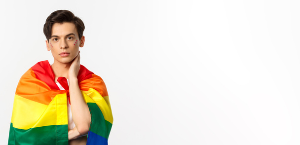 Μια φωτογραφία ενός όμορφου γκέι άντρα με γκλίτερ στο πρόσωπο, τυλίγεται με τη σημαία του ουράνιου τόξου και αγγίζει απαλά το πρόσωπο, κοιτάζοντας κάμερα, λευκό φόντο. - Φωτογραφία, εικόνα