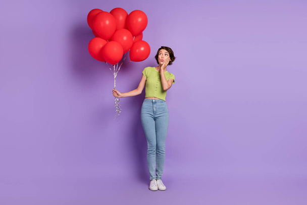 Photo of impressed attractive young woman dressed green t-shirt looking red air balls hand arm cheek cheekbone isolated purple color background. - Foto, Imagem