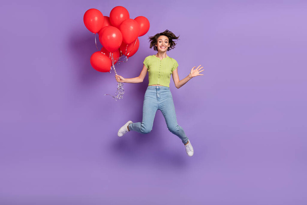 Zdjęcie pięknej funky młoda dama nosić zielony t-shirt uśmiechnięty skoków wysoki gospodarstwa czerwone balony odizolowane fioletowy kolor tła. - Zdjęcie, obraz