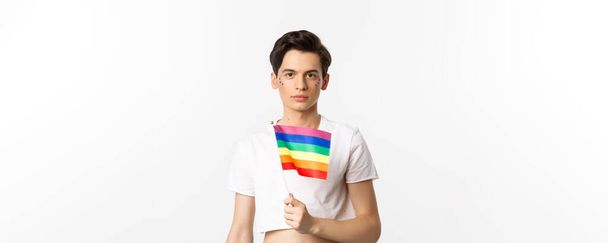 Pride and lgbtq concept. Waist up shot of attractive anrogynous man holding rainbow flag, having glitter on face and looking at camera, standing over white background. - Photo, Image