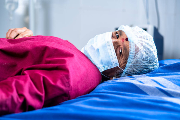 Starší nemocný pacient na nemocničním lůžku s maskou a operačním pláštěm při pohledu do kamery - koncept lékařské péče, nemoci a uzdravení. - Fotografie, Obrázek