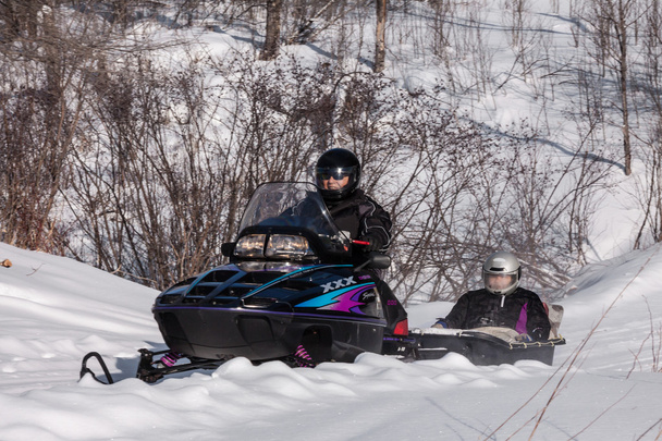 Snowmobile transporte duas pessoas no inverno, um dirigindo uma moto de neve, o outro descansando no trailer
 - Foto, Imagem