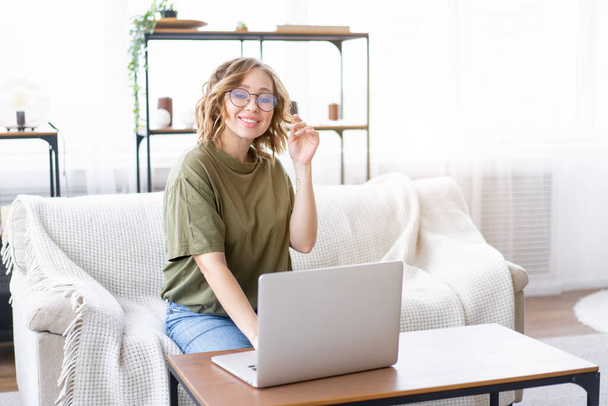 Woman with glasses use laptop typing keyboard sitting couch big window background home interior Freelance female working from home Distance learning student relaxing watch lessons video conferenc - 写真・画像