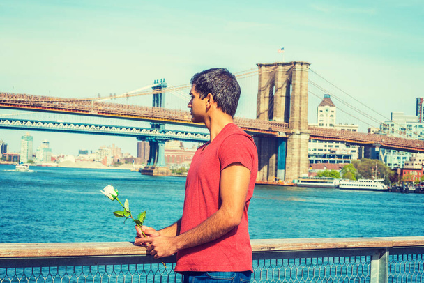 Я скучаю по тебе, жду тебя. Ост-Индский американец в красной футболке, стоящий у реки, держащий белую розу, отворачивающийся. Манхэттен, Бруклинские мосты на заднем плане.  - Фото, изображение
