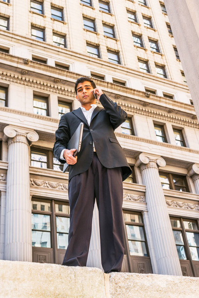 Ostindischer amerikanischer Geschäftsmann auf Reisen, der in New York arbeitet. Im schwarzen Anzug und mit Laptop steht ein junger Mann auf einer Straße mit hohen Gebäuden und telefoniert. - Foto, Bild