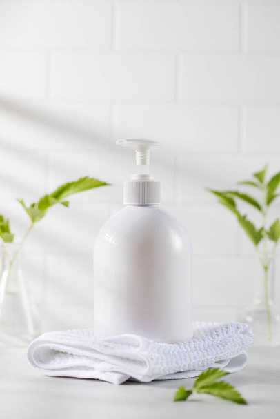 White plastic dispenser with a cosmetic product on a background of white tiles and green plants with a place for your logo. Skin care. - Fotó, kép