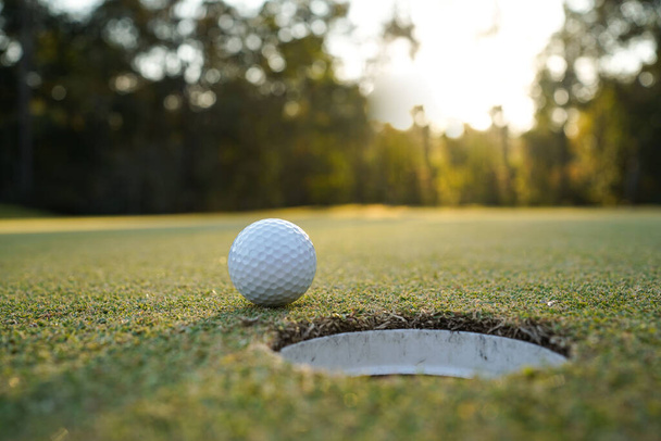 Golf ball on green grass in the evening golf course with sunshine background. Golf ball on the edge of hole on the green grass with warm tone and sunset.                                - Foto, afbeelding